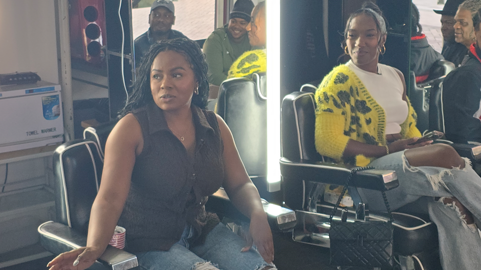 The two women are sat on barber chairs at the barbers. Chanelle is wearing a black sleeveless top and jeans. Talissa is wearing a yellow cardigan over a white top with jeans. Several men are in the background.
