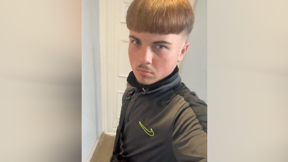 Aidan Tottman is taking a selfie with brown hair brushed to the front of his face and shaved around the side of his head. He is wearing a Nike jumper and is stood in front of a white door