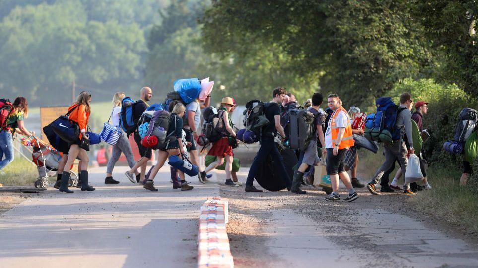 A group of people carrying tents and sleeping bags across a road while a steward wearing an orange hi-vis directs them through a gap in the hedge.