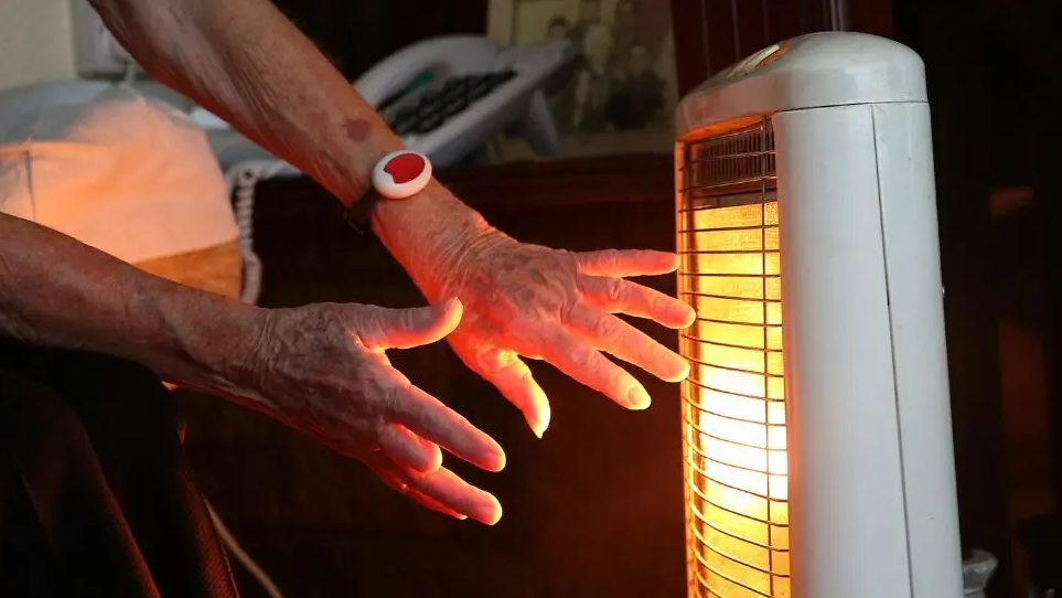 An elderly person warming their hands by a bar fire. The fire is in a white plastic case and has a silver grill on the front. There is an orange glow from inside the grill. Only the person's hands are visible and they are holding both towards the body of the heater.