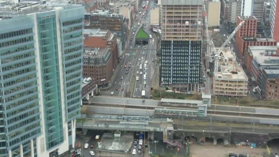 Aerial shot of city from a drone. There are lanes of vehicles and a bridge over them. There are also tall buildings and a number of smaller buildings and a crane in the picture. 