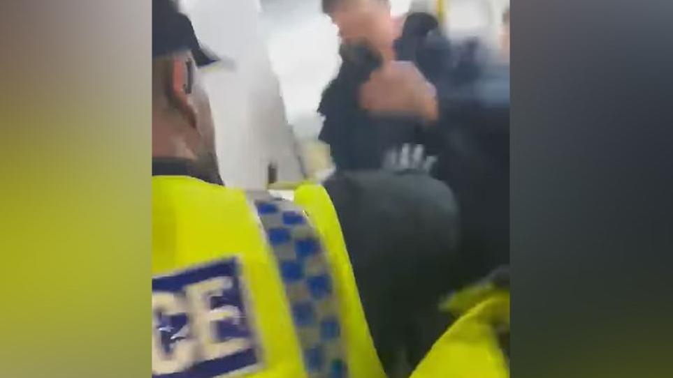 Police officer in hi-vi vest putting his hand out as a male youth with his face blurred stands on a train at Manchester Victoria holding his mobile phone up