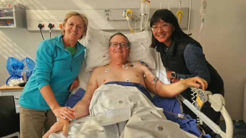 Roland Cherry is lying in a hospital bed with his wife and another woman by his side. He has tube and stickers on his bare chest with the two women either of him. His wife is wearing a blue top and brown trousers. The second woman is in dark top and trousers. All are smiling in the photo.
