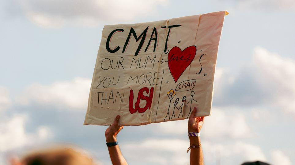 A fan holds a handmade sign in the air reading "CMAT, our mum loves you more than us!". There are also drawings of a heart and a family. 