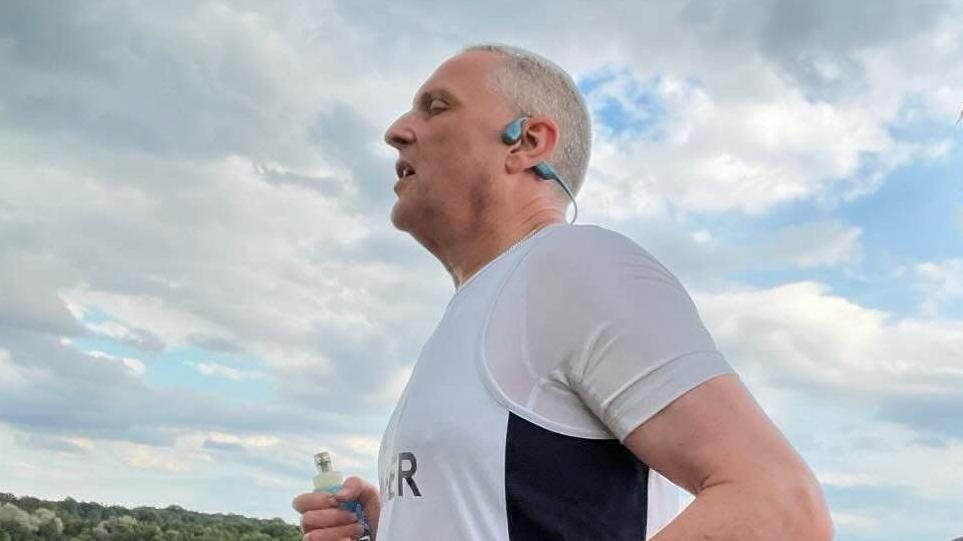 A profile image of Mr Goodall taken while he is running, showing the top half of his torso and upper arm. He is wearing a white T-shirt with a white vest over it that has a black stripe going down the side of it. He is wearing grey earphones and his eyes are closed. His right arm can be seen at the bottom of the image frame clutching what appears to be an electrolytes tube. 