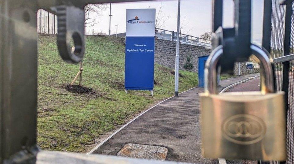 A sign that reads: "Driver & Vehicle Agency Welcome to Hydebank Test Centre" behind a padlocked gate