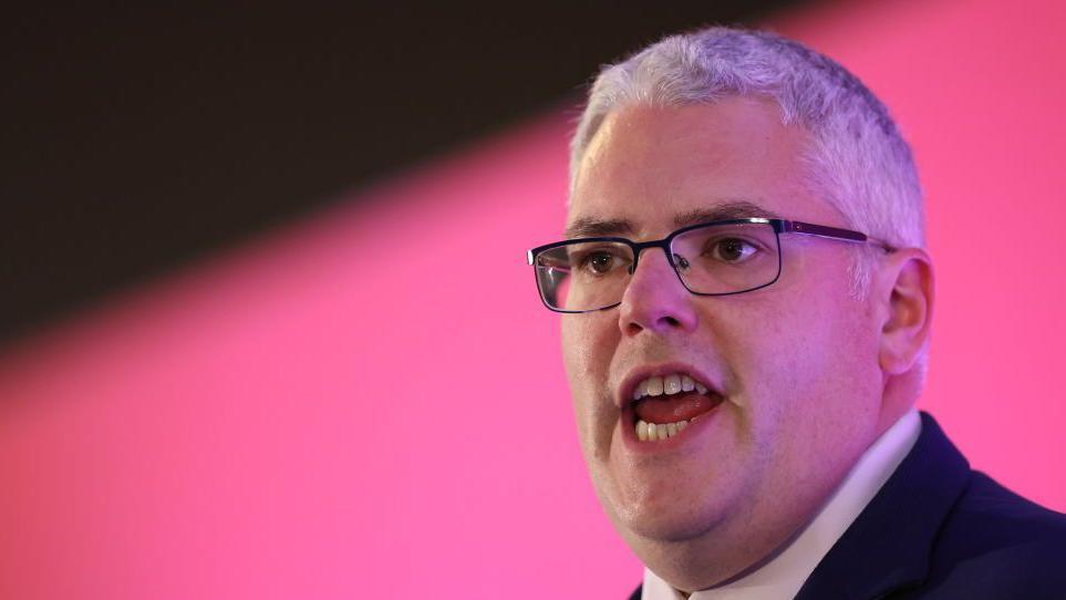Gavin Robinson has short grey hair and is wearing black rectangular metal glasses. He is wearing a suit jacket with a white shirt and is talking in the image. The background behind him is pink and blurred.
