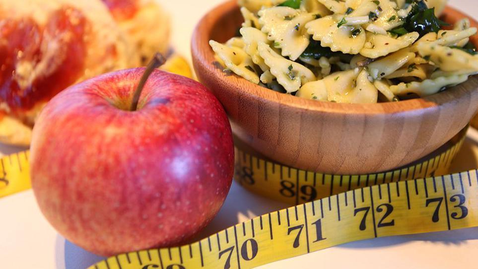 An illustrative image showing "healthy food". A shiny red apple is next to a small wooden dish of pasta. A yellow tape measure winds around them.