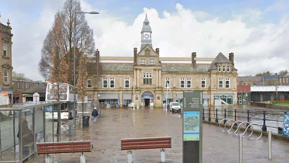 Street view of Darwen Market, a large stone building with a clock tower in the middle, with a large flagged square in front 