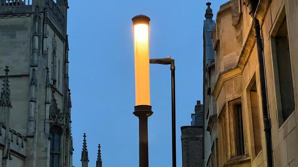 A lamp lit up against the evening sky. It is tall and thin and there are ornate university buildings on either side