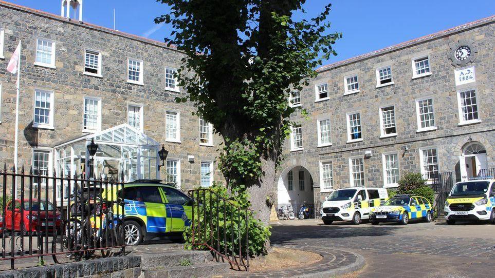 Guernsey Police headquarters. A grey building with three floors and windows on eat floor. There are police vans and cars parked in the car park. There is a tree in the middle of the car park. The sky is blue. 
