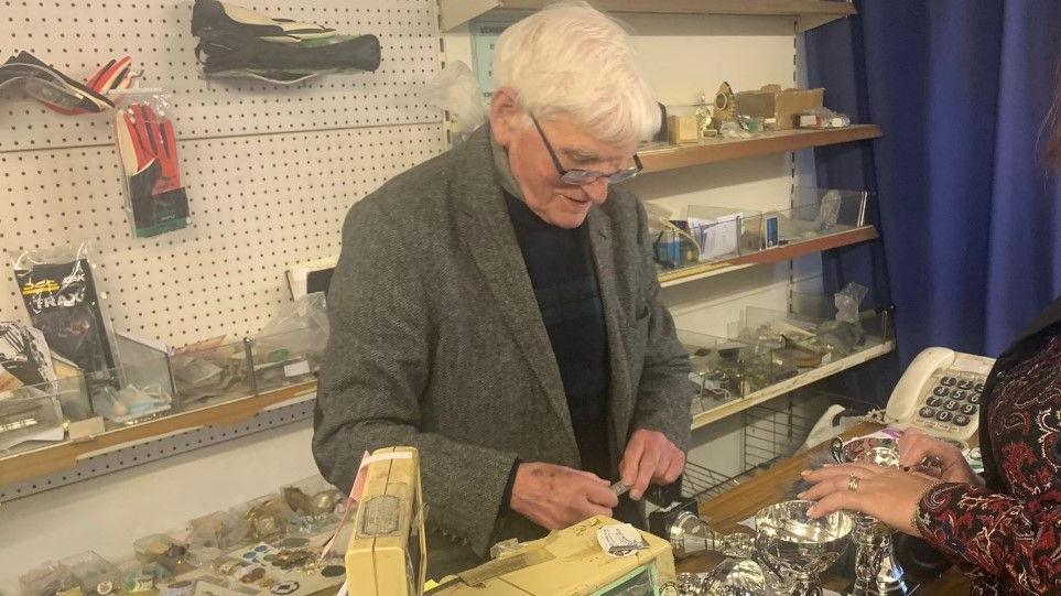 John 'Jobby' Crossan pictured behind the counter of his shop. 
A number of small trophies are in front of him and he is facing a customer. 
The female customer is wearing jewellery and is in a patterned top. 