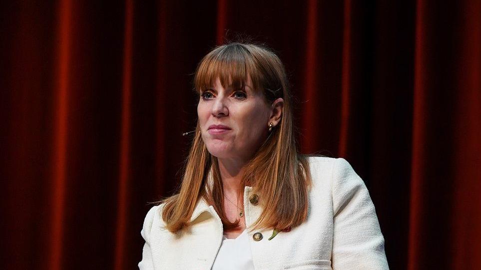 Angela Rayner wearing a white jacket sitting down against a red curtain backdrop