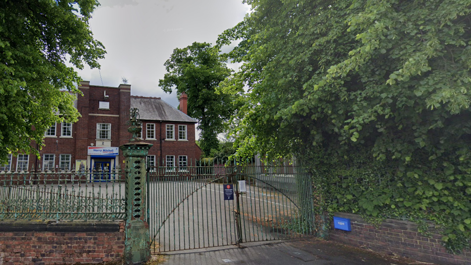 An exterior shot of Harry Mitchell leisure centre in Smethwick. Metal gates lead into a car park, on the other side of which is a large brick building with a sign above the entrance reading "Harry Mitchell".