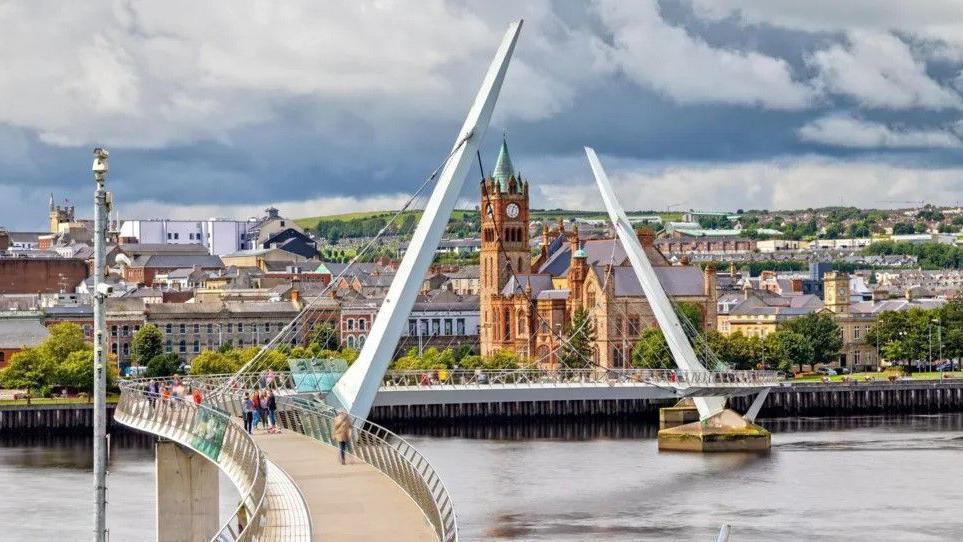 A view of Londonderry with the Peace Bridge over the Foyle river in the foreground and the Guildhall and other building in the background.
