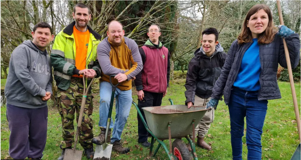 Five men and a woman standing in a garden holding gardening tools. There is a wheelbarrow next to them