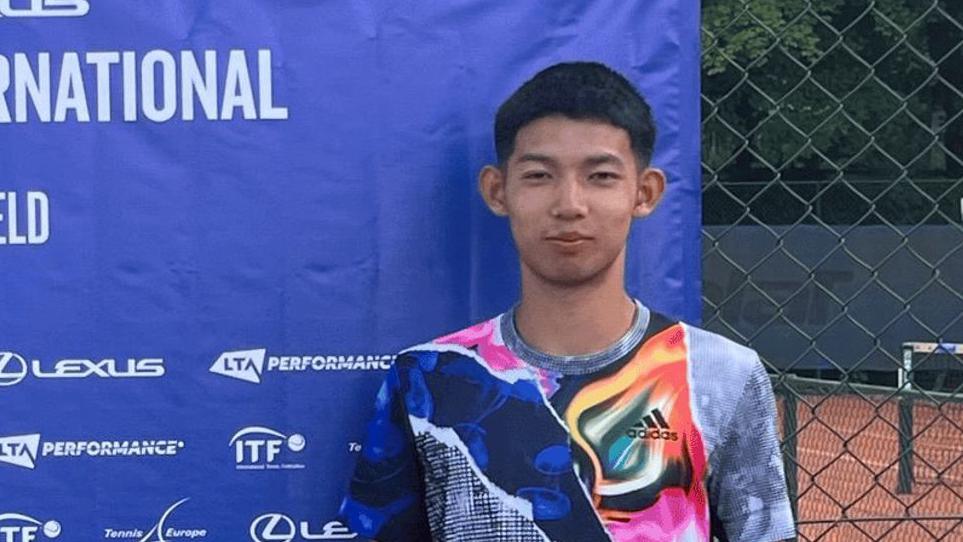 A young man with short dark hair and a multi-coloured sports top standing in front of a blue advertising screen with a clay tennis court partially visible behind him
