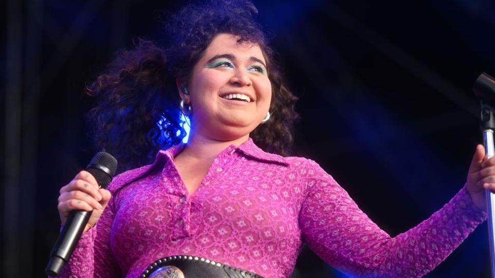 Remi Wolf on stage. Remi has curly brown hair and bright blue eye make-up. She wears a patterned pink jumpsuit and smiles into the crowd. 