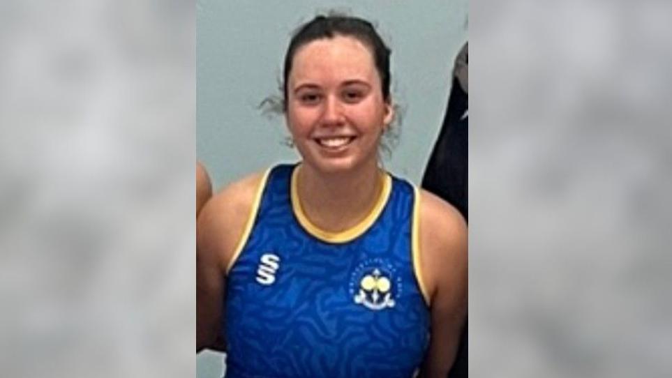 Hannah Cliftlands, a young women with brown hair in a ponytail wearing a blue and yellow netball kit smiling at the camera. 