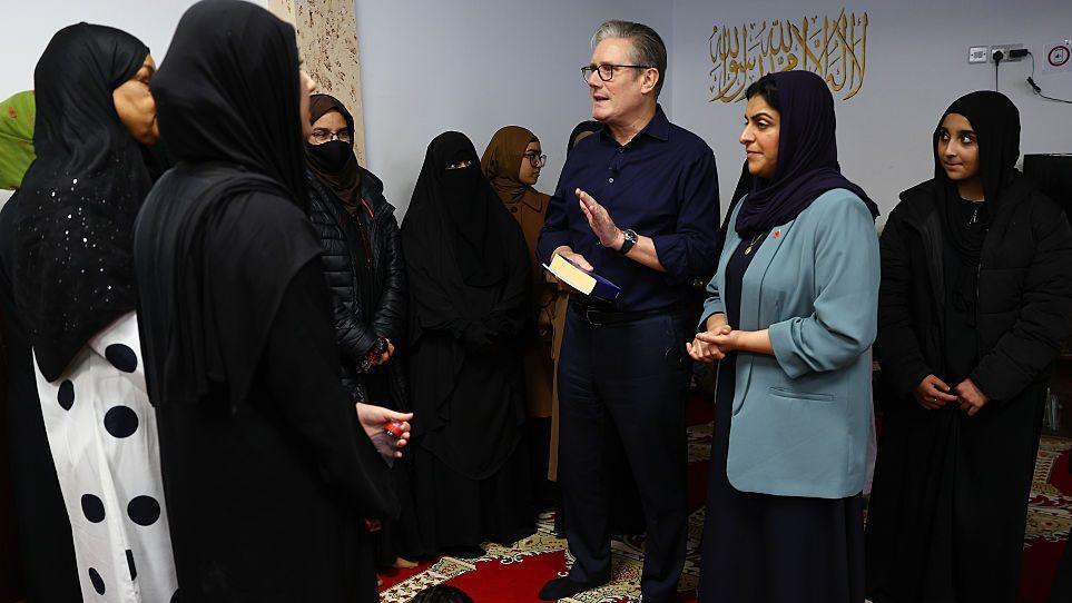 A man wearing glasses is standing next to a woman, wearing a cadet blue suit and a headscarf over her head. They are talking to a group of people.
