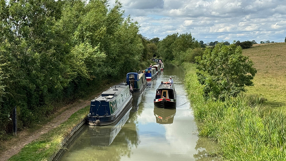 An elevated view of a canal in Hillmorton, probably taken from a bridge, with a line of at least five narrowboats moored up on the left hand side and a single narrowboat, passing them, approaching the photographer. A towpath fringed with trees is on the left and a green field is on the right of the canal