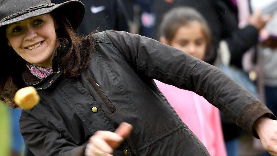 A woman wearing a black coat and a black hat throws a small round scone-like biscuit. She is smiling.