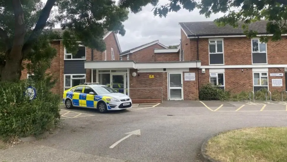 A police car parked outside a red-bricked building. The building has a car park to its front.
