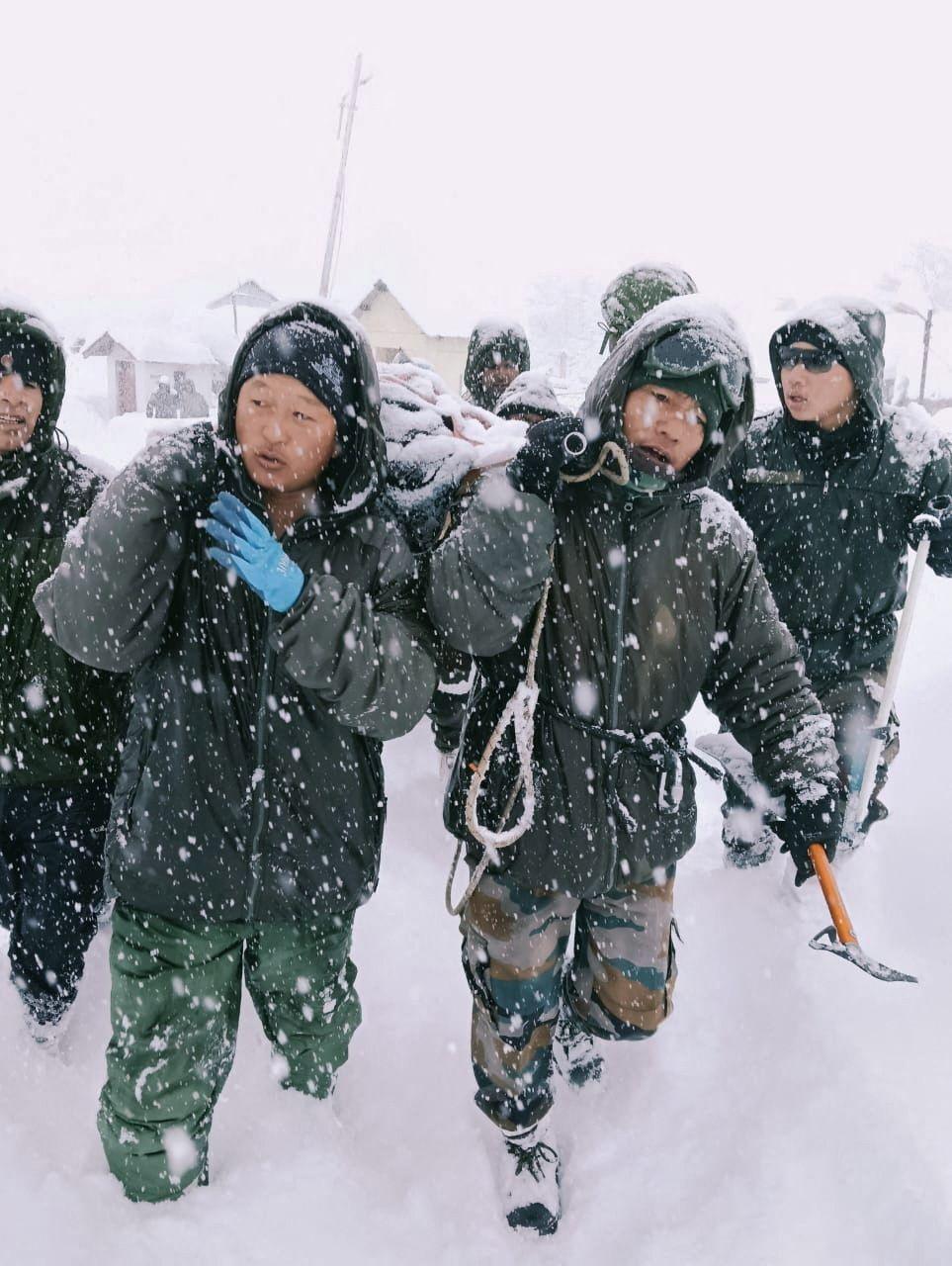 Two rescuers from the Indian Army, dressed in green overalls, carry items over their shoulders as they trek through heavy snow
