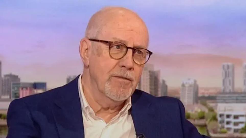 Colin Parry wearing a white shirt, navy blazer and glasses on the BBC Breakfast studio with a view of Salford in the background