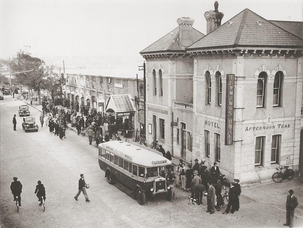 A black and white image of a bus on a busy early 20th Century street, with shops and a hotel behind it.