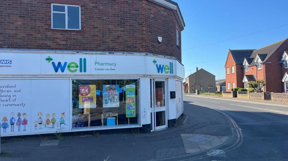 The Well Pharmacy on the junction of High Street and Beach Road in Caister. The building dates from the 1950s and has Crittall windows on the upper storey, and a façade with large windows covered in a vinyl depicting families in a child-like drawing. Houses on High Street can be seen on the right of the image.