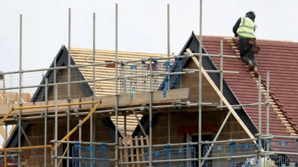 A builder in a hi-vis jacket, is working on a roof of a newly built house. Scaffolding surrounds a row of houses.