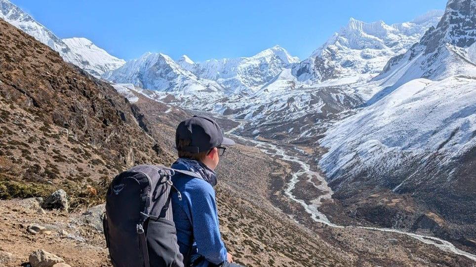 Rio sat looking over the Himalayan mountains. He has a rucksack on, with a blue jacket on and a blue cap.