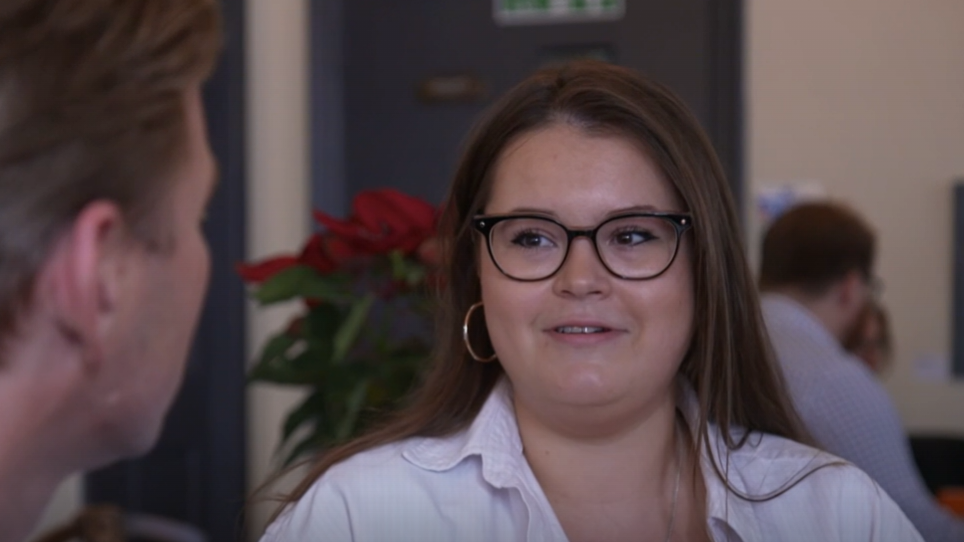 Jordan Till is looking at a man on her left who she is having a conversation with. She wears a white longs sleeved shirt, has hoop earrings and long brown hair. She also wears a silver necklace and circle framed black glasses.