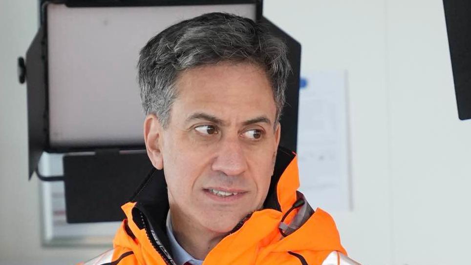 Ed Miliband looks away from the camera. He has dark short hair with some grey in it. He is wearing a bright orange coat with a shirt just visible underneath