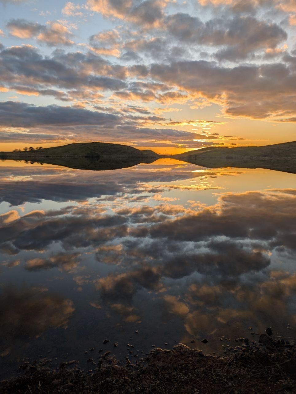 A loch with clouds above it as the sun sets. The clouds are also reflected on the water.