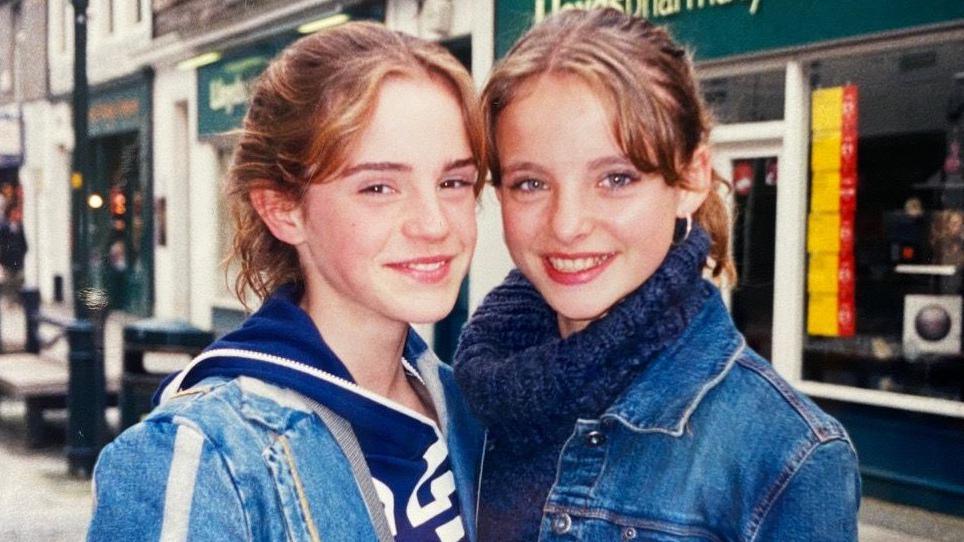 Two individuals stand close together on a street, both wearing denim jackets, in front of a pharmacy