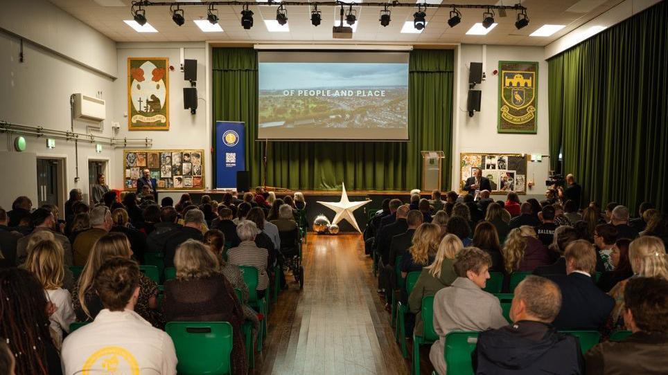 People have filled the seats in the assembly hall at Alsop High School. They are all looking towards a large screen showing the documentary.