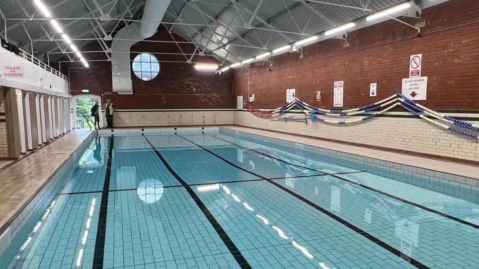 A large swimming pool inside a brick building with a pointed metal roof and changing rooms to the left hand side