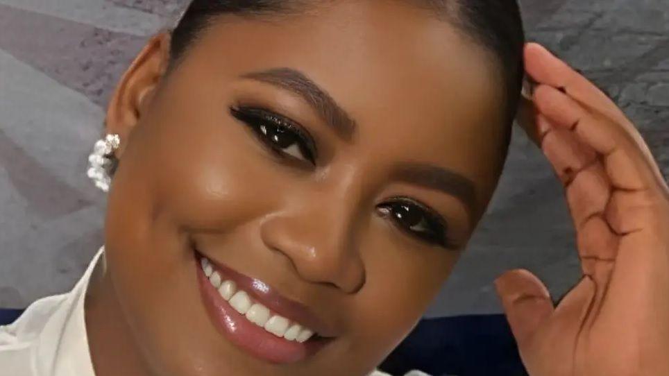 A woman with dark hair smiles for the camera as she holds a hand to her head. She is wearing a white blouse and white earring.