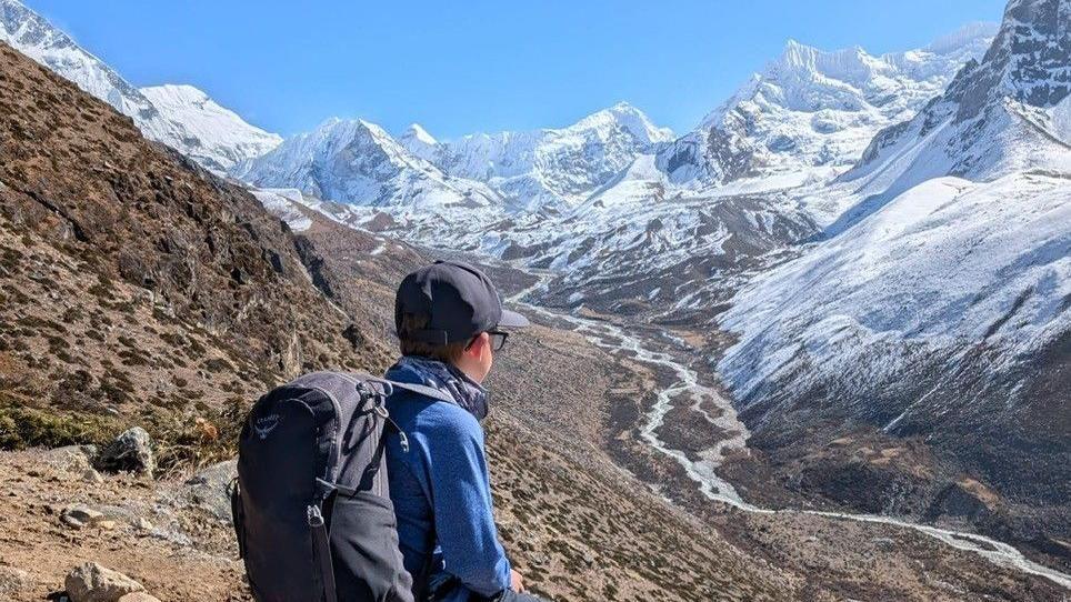 Rio looking over the Himalayan mountains. He has a rucksack on, with a blue jacket on and a blue cap.