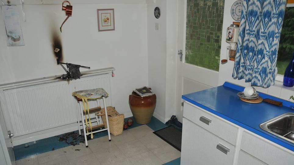 Photograph of Una Crown's kitchen, which has white walls, white counters with a blue countertop, blue and white curtains, white back door and a burn mark on the wall around a towel rail and radiator