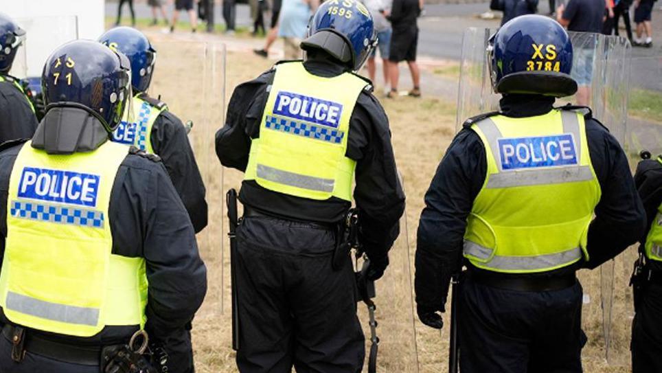 Generic image of the backs of four riot police wearing helmets and high visibility jackets, watching a crowd of people in the distance