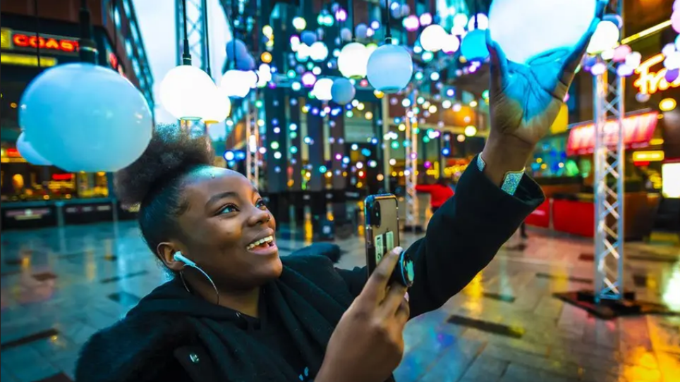 A woman holding a hanging light-bulb which is part of an illuminated art installation and taking a picture with her phone.