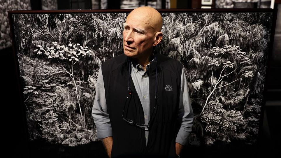 Sebastião Salgado standing in front of a black and white photo of trees in the Amazon