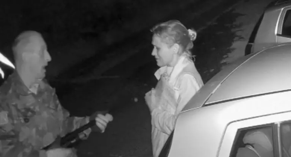 Black and white CCTV image of a man in a camouflage jacket holding a shotgun to the chest of a scared looking woman in a light coat, They are stood on a road near some parked cars 