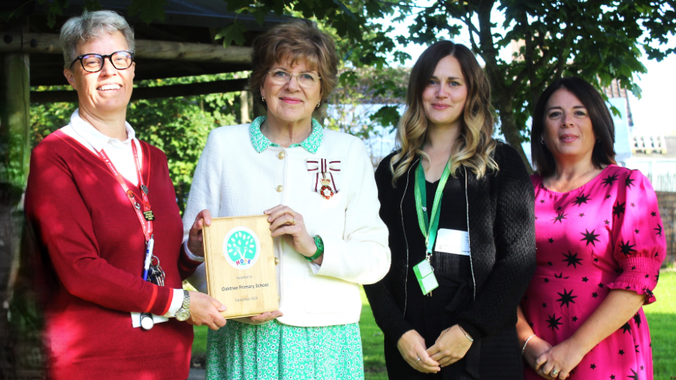 Four women stood in a garden smiling at the camera. Two of the women are holding a Hope Award certificate, which has the name of the school - Oaktree Primary School - on it.