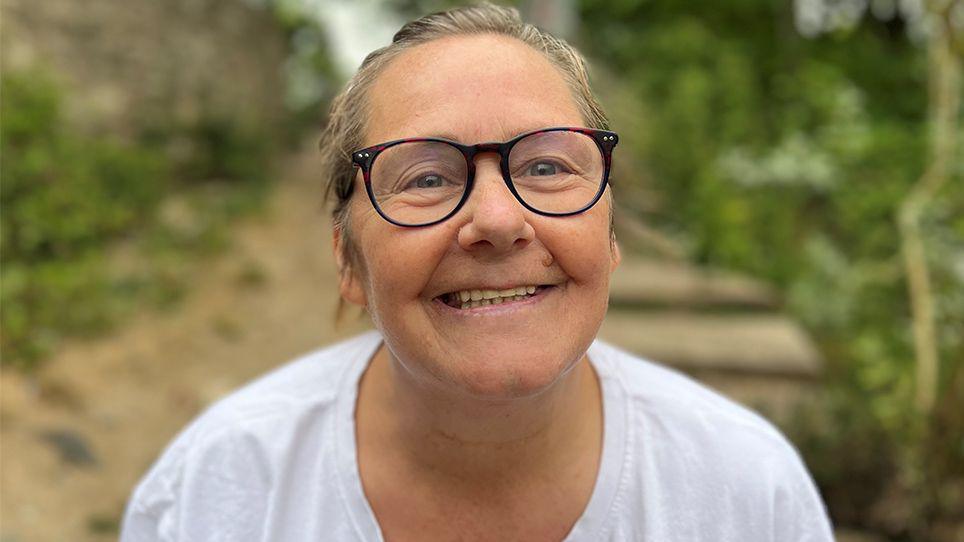 Head and shoulders image of Viv Storey. She is standing outside with greenery behind her. She is smiling, wearing glasses and a white t-shirt and her hair is tied back off her face