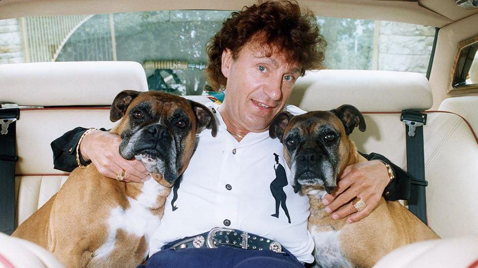 Frank Pearson with brown curly hair wearing a white shirt and denims hugging two boxer dogs with each arm in the back of a car 