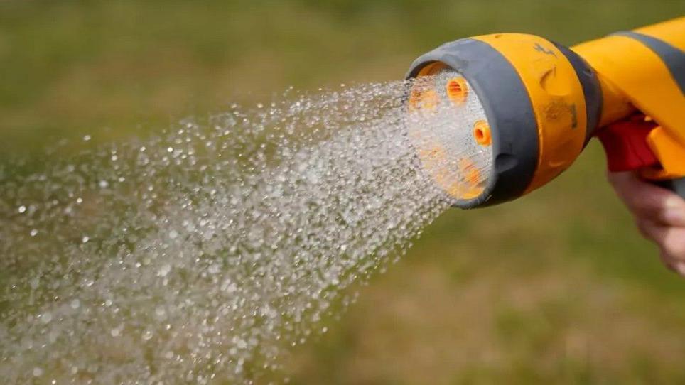 A person holding the nozzle of a yellow hosepipe with water gushing out of it. It is being sprayed onto some grass. 